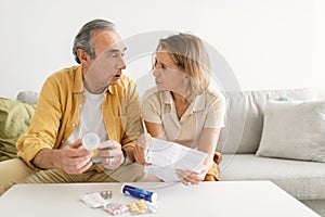 Senior spouses reading leaflet of drugs before drinking pills, sitting on sofa in living room and looking at each other