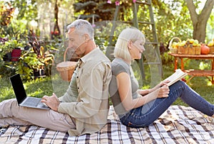 Senior spouses resting in garden, man using laptop computer, woman reading book, sitting back to back, side view