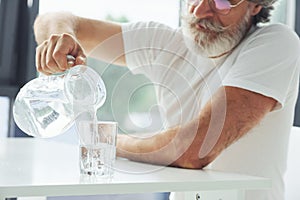 Senior stylish modern man with grey hair and beard indoors pouring water into glass