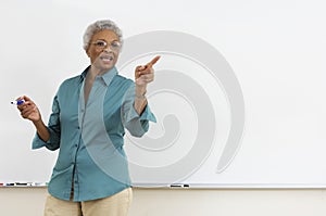 Senior teacher pointing while gesturing against white board in classroom