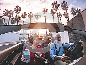 Senior trendy couple inside a convertible car on holiday time - Mature people having fun doing a road trip during vacation -