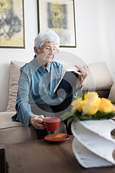 Senior white-haired woman reading a book and drinking coffee at home