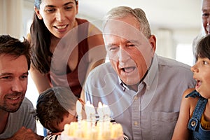 Senior white man celebrating with his family blowing out the candles on birthday cake, close up