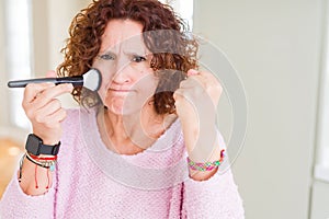 Senior woman applying blush on face using a brush annoyed and frustrated shouting with anger, crazy and yelling with raised hand,