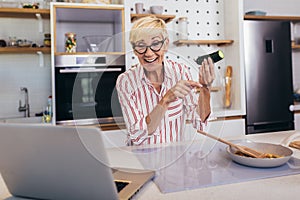 Senior woman in apron and with eyeglasses standing in kitchen, using laptop and preparing healthy meal