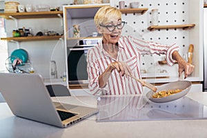 Senior woman in apron and with eyeglasses standing in kitchen, using laptop and preparing healthy meal