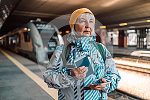 Senior woman with backpack in yellow hat holds biometric passport and train ticket and using smartphone waiting train on