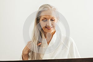 Senior woman in bathrobe looking down while adjusting her long white hair