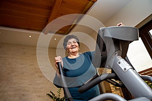 A senior woman with black short hair in a turquoise T-shirt is practicing on a Nordic ski simulator. She is cheerful and laughs