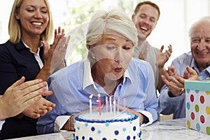 Senior Woman Blows Out Birthday Cake Candles At Family Party