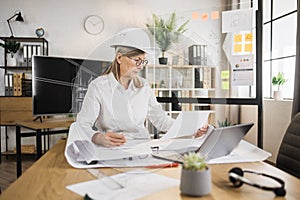 Senior woman builder working on laptop while looking on papers or sketches.