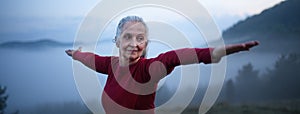 Senior woman doing breathing exercise in nature on early morning with fog and mountains in background.