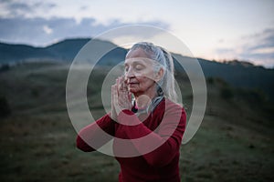 Senior woman doing breathing exercise in nature on early morning with fog and mountains in background.