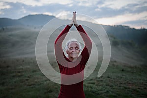 Senior woman doing breathing exercise in nature on early morning with fog and mountains in background.