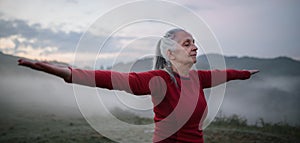 Senior woman doing breathing exercise in nature on early morning with fog and mountains in background.