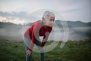 Senior woman doing breathing exercise in nature on early morning with fog and mountains in background.