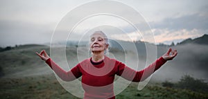 Senior woman doing breathing exercise in nature on early morning with fog and mountains in background.