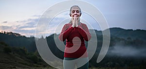 Senior woman doing breathing exercise in nature on early morning with fog and mountains in background.