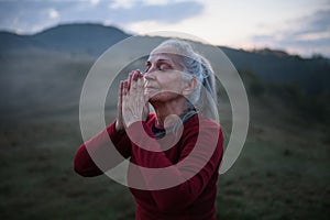 Senior woman doing breathing exercise in nature on early morning with fog and mountains in background.