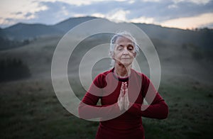 Senior woman doing breathing exercise in nature on early morning with fog and mountains in background.