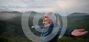 Senior woman doing breathing exercise in nature on early morning with fog and mountains in background.