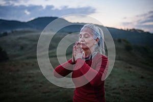 Senior woman doing breathing exercise in nature on early morning with fog and mountains in background.