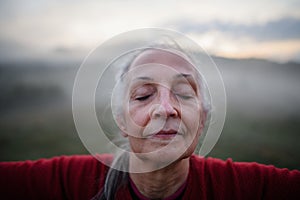 Senior woman doing breathing exercise in nature on early morning with fog and mountains in background, close-up