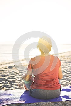 Senior woman doing exercise on yoga mat at the beach, meditating. Shot from back