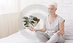 Senior woman doing yoga, meditating in lotus position on bed at home