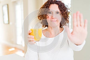 Senior woman driking a glass of fresh orange juice with open hand doing stop sign with serious and confident expression, defense