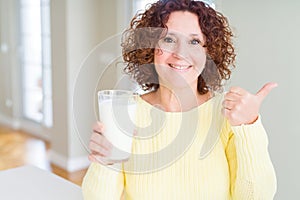 Senior woman drinking a glass of fresh milk pointing and showing with thumb up to the side with happy face smiling