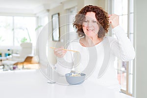 Senior woman eating asian noodles using chopsticks annoyed and frustrated shouting with anger, crazy and yelling with raised hand,