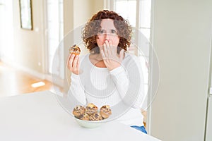 Senior woman eating chocolate chips muffins cover mouth with hand shocked with shame for mistake, expression of fear, scared in