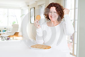 Senior woman eating healthy whole grain biscuit at home annoyed and frustrated shouting with anger, crazy and yelling with raised