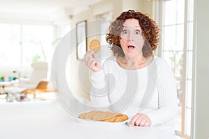 Senior woman eating healthy whole grain biscuit at home scared in shock with a surprise face, afraid and excited with fear