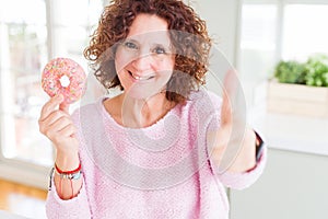 Senior woman eating pink sugar donut happy with big smile doing ok sign, thumb up with fingers, excellent sign