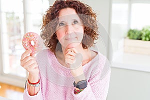 Senior woman eating pink sugar donut serious face thinking about question, very confused idea