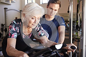 Senior Woman Exercising On Cycling Machine Being Encouraged By Male Personal Trainer In Gym