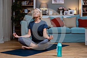 Senior woman exercising while sitting in lotus position. Active mature woman doing stretching exercise in living room at home. Fit