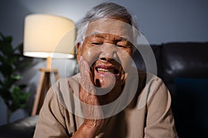 senior woman feels strong toothache while sitting on sofa in the living room at night