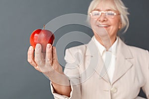 Senior woman in fromal suit and glasses studio standing isolated on gray holding apple close-up blurred smiling joyful