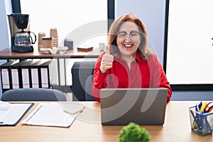 Senior woman with glasses working at the office with laptop smiling happy and positive, thumb up doing excellent and approval sign