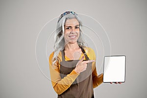 Senior woman with gray hair, in apron, holding tablet, offering farm product recommendations