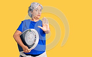 Senior woman with gray hair holding weight machine to balance weight loss with open hand doing stop sign with serious and