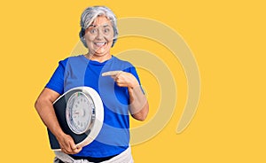 Senior woman with gray hair holding weight machine to balance weight loss smiling happy pointing with hand and finger