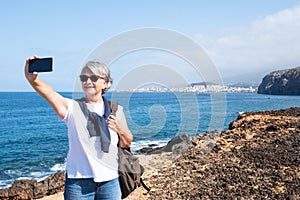 Senior woman gray hair takes a selfie on the cliff with a beautiful seascape. Adult 70 years old. Blue background. One people