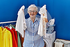 Senior woman with grey hair holding clean white t shirt and t shirt with dirty stain smiling with a happy and cool smile on face