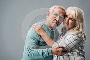 senior woman with grey hair hugging upset husband on grey.