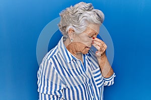 Senior woman with grey hair standing over blue background tired rubbing nose and eyes feeling fatigue and headache