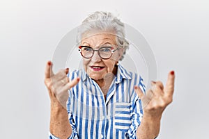 Senior woman with grey hair standing over white background shouting with crazy expression doing rock symbol with hands up